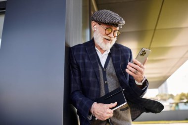 Son sınıf öğrencisi şık bir binaya yaslanmış, şık bir kıyafet giyerken telefonunu kontrol ediyor..