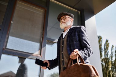 İyi giyimli bir son sınıf öğrencisi, sonbaharda çağdaş bir binanın yanında dolaşırken bir çanta ve telefon taşır..