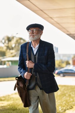 Şık ceketli ve düz şapkalı son sınıf öğrencisi sonbaharda modern binalar tarafından zarif bir şekilde yürür..