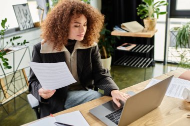 Kadın, şık bir ofis ortamında dizüstü bilgisayarı üzerinde çalışırken özenle evrakları inceliyor..