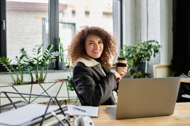 A woman smiles and holds a coffee cup while working at her laptop in a bright room. clipart