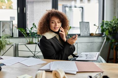 A woman with curly hair talks on the phone while focused on her laptop in a bright room. clipart