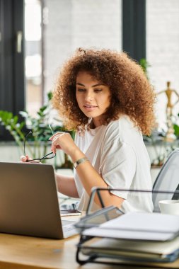 A young woman studies her laptop intently, adjusting her glasses in a vibrant workspace. clipart