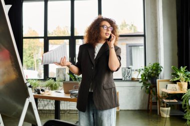 The woman engages in a lively discussion on her phone while holding papers in her office. clipart