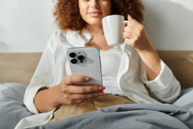 Rahat bir kadın sabah saatlerinde kahvesini yudumlar ve telefonunu rahatça yatağına kaydırır..