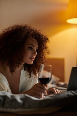 In a cozy bedroom, a woman enjoys a glass of red wine while focusing on her laptop. clipart