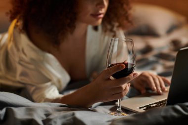 A woman enjoys a glass of wine while working on her laptop in a comfortable bedroom setting. clipart