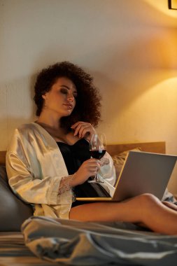 Relaxing in a serene bedroom, a woman sips wine while engaging with her laptop. clipart