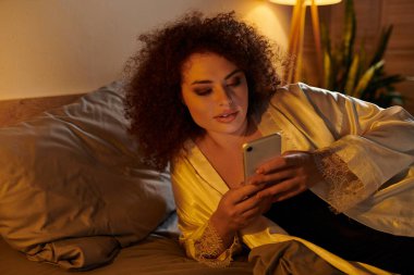 Saten bornozlu bir kadın gece çökerken yatakta dinleniyor, akıllı telefonuna dalmış..