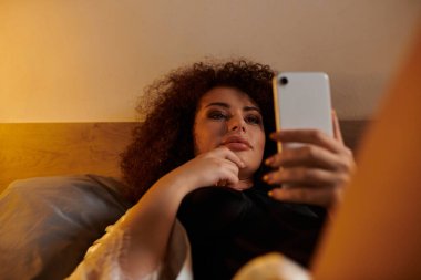 Yatakta dinlenen bir kadın, akıllı telefonuyla düşüncelere dalmış..
