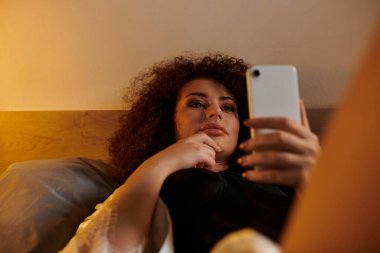 Relaxing in bed, a woman scrolls through her smartphone, lost in thought. clipart