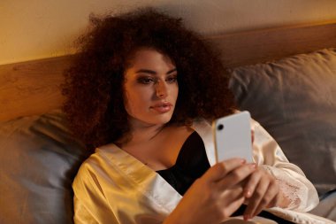 A woman with curly hair enjoys time in bed, absorbed in her smartphone under warm lighting. clipart