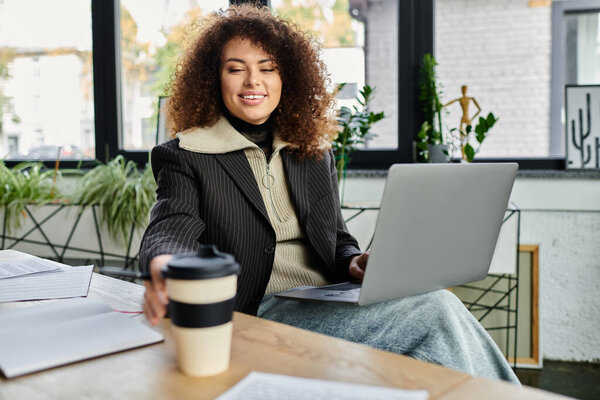 Женщина с вьющимися волосами улыбается, когда тянется за чашкой кофе во время работы.