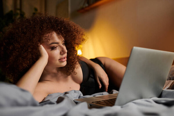 Женщина в интимной одежде лежит на кровати, используя свой ноутбук в мягком вечернем свете.