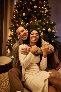 A loving couple shares a warm embrace at home, surrounded by festive decorations during Christmas. clipart