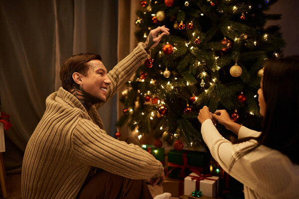 A loving couple shares laughter while adorning their Christmas tree with festive ornaments indoors.
