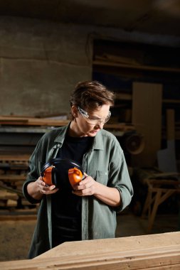 The carpenter prepares her hearing protection while surrounded by wooden materials and tools in her workshop. clipart