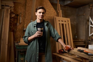 A skilled carpenter takes a moment to savor her coffee while surrounded by tools and wood in the workshop. clipart