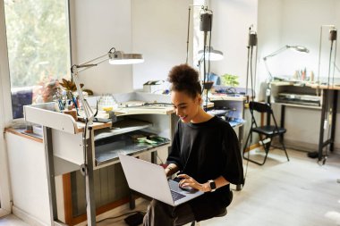 The woman smiles while typing on her laptop, surrounded by tools and elegant jewels. clipart