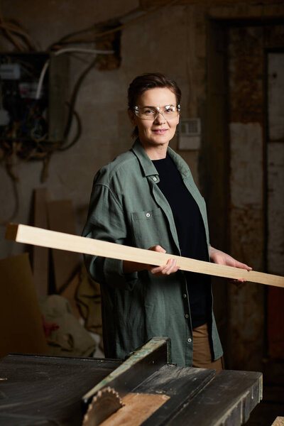 A skilled female carpenter holds a wooden beam, showcasing her craftsmanship in a vibrant workshop.
