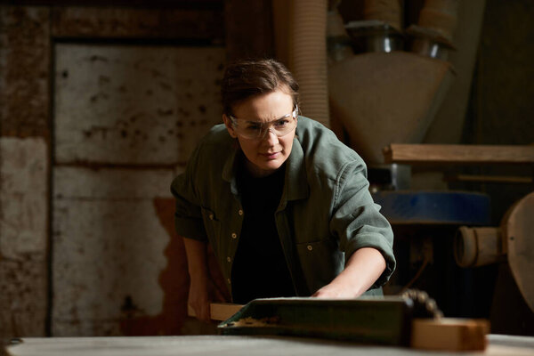 A dedicated female carpenter concentrates on her wood project in a lively workshop filled with tools.