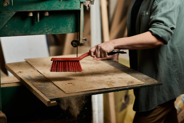 A skilled carpenter cleans up wood shavings with a broom, focused on her craft in a lively workshop environment.