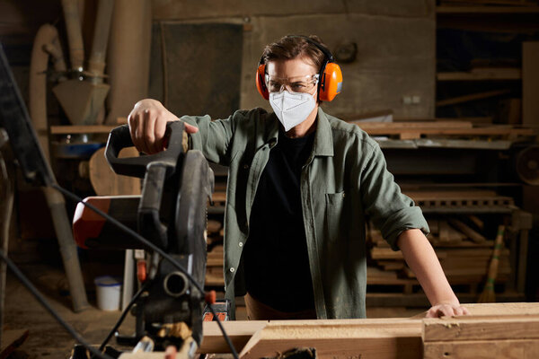 A focused carpenter expertly maneuvers machinery, cutting wood while wearing protective gear in her workshop.