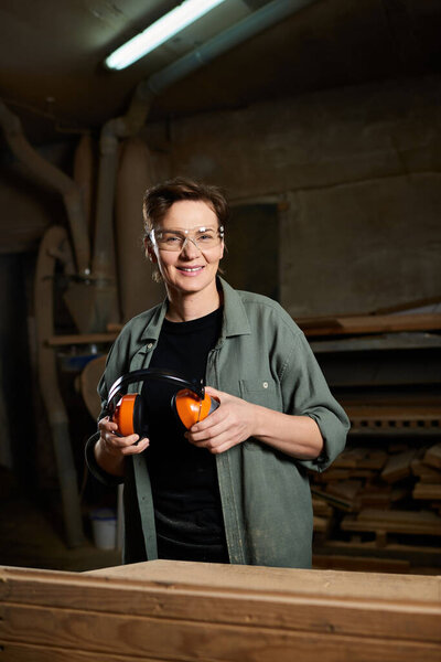 In a well-lit workshop, a female carpenter holds her ear protection, ready to craft her next woodworking project.