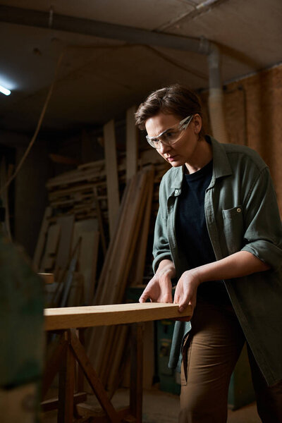 Целеустремленный плотник женского пола фокусируется на своей работе, мастерски формируя дерево в шумном помещении мастерской.