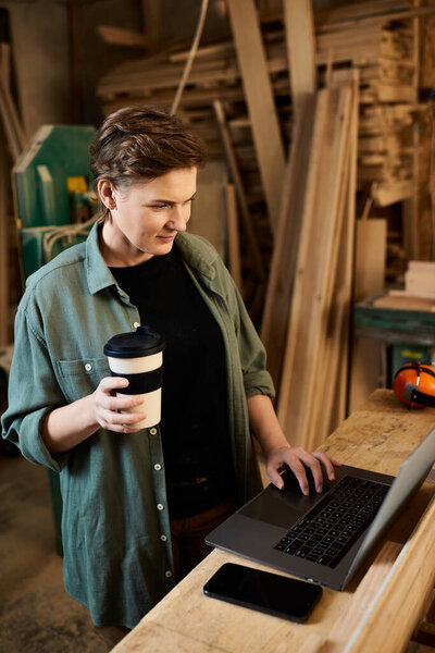 A dedicated carpenter reviews plans on her laptop, coffee in hand, within a vibrant woodworking workshop.