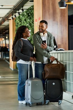 A joyful young couple with luggage smiles at each other in a stylish hotel lobby, ready for adventure. clipart