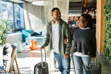 Genç bir Afrikalı Amerikalı çift, bavullarıyla canlı otel lobisinde yürürken gülümsüyor..