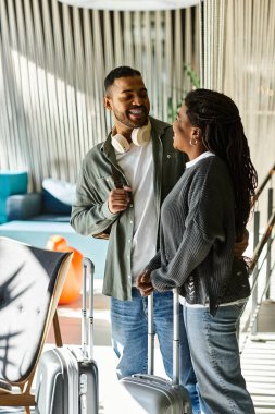 A young African American couple smiles and shares a moment in a hotel lobby before their vacation begins. clipart