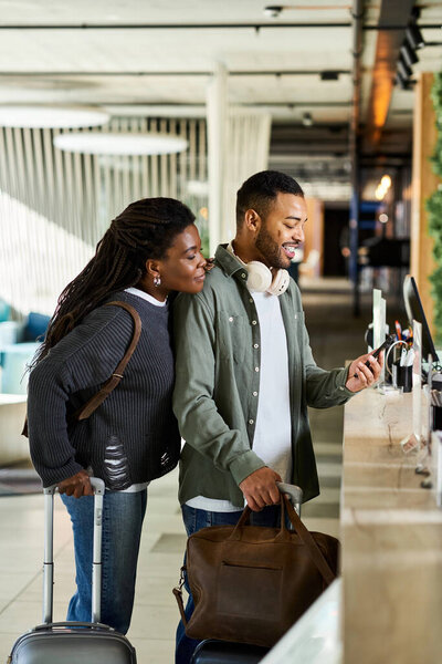 Молодая афроамериканская пара с багажом радостно регистрируется в оживленном вестибюле отеля вместе.