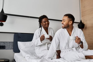 A joyous couple shares smiles and warm drinks while relaxing in their hotel room on vacation. clipart