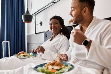 Genç bir çift otel odasında dinleniyor, keyifli bir yemek yiyor ve gülümsüyor, birlikte tatillerinin tadını çıkarıyorlar..