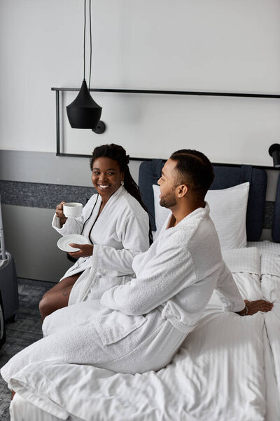 A joyful young couple relaxes in luxurious robes, sharing sweet moments while enjoying breakfast in bed.