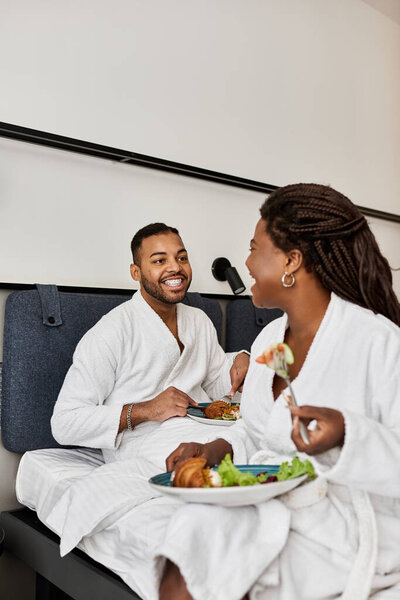 A young couple shares laughter and delicious food while relaxing together in their hotel room.