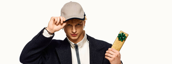 В праздничной атмосфере, модный человек надевает модный наряд и кепку, представляя красиво завернутый подарок с бантиком. Его уверенная поза захватывает дух Рождества 2025 года.