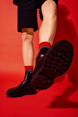 Gösterişli siyah çizmeler, canlı kırmızı çoraplar ve çarpıcı bir kırmızı zemine karşı kendine güvenen pozlar sergileyen bir adam moda tatil modası sergiliyor. Bu tarz 2025 Noel ruhunu yansıtır..