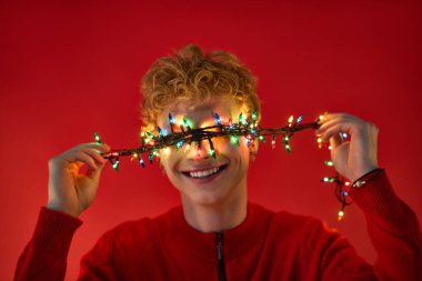 Canlı kırmızı bir ortamda, genç bir adam neşeyle ışıldıyor, neşeyle gözlerinde renkli Noel ışıkları tutuyor..