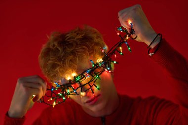 Bir adam renkli Noel ışıklarını yüzüne canlı kırmızı bir arka plan boyunca şakacı bir şekilde örterek yaratıcı tatil modasını sergiliyor. Neşeli ifadesi 2025 'in neşeli ruhunu yansıtıyor..
