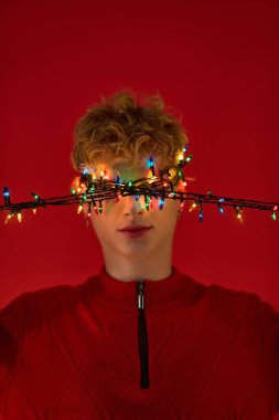 Brightly lit Christmas lights drape across a mans face, embodying the holiday spirit in a striking red setting. His joyful expression showcases his stylish seasonal outfit, perfect for celebrations. clipart