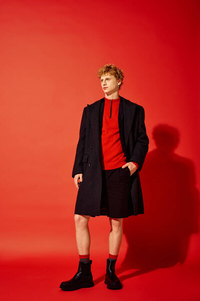 Стильный мужчина демонстрирует праздничную моду с уютным красным топом и элегантным черным пальто. Он уверенно стоит на ярком красном фоне, вызывая праздничный дух в этом рождественском сезоне в 2025 году..