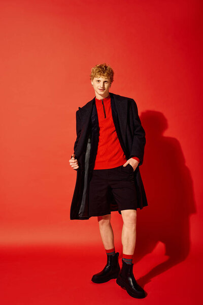 Молодой человек демонстрирует яркий праздничный наряд с красным топом, черным пальто и стильными шортами. Праздничная обстановка излучает дух Рождества на ярко-красном фоне.