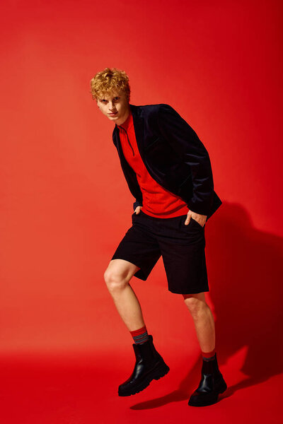 Стильный мужчина демонстрирует смелую праздничную моду в яркой красной обстановке, одетый в яркий ансамбль красного топа и шорты в паре с черными сапогами. Этот взгляд переопределяет праздничный наряд на 2025 год.