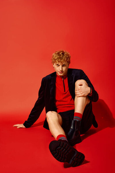 Модный человек сидит на ярком красном фоне, выражая уверенность в своем праздничном ансамбле. Его наряд отличается яркими цветами и уникальными узорами, идеально подходит для сезонных праздников в 2025 году.