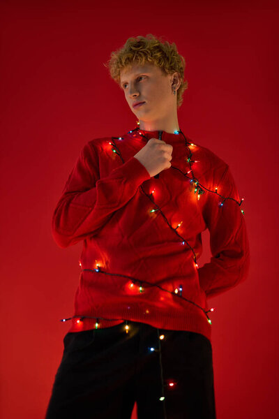 Стильный молодой человек завернут в яркие рождественские огни на ярком красном фоне. Его праздничный красный свитер усиливает радостную атмосферу, прекрасно улавливая праздничный дух 2025 года.