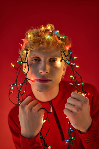 Стильный молодой человек позирует на ярко-красном фоне, игриво переплетенном в красочные рождественские огни. Его радостное выражение лица отражает праздничный дух курортного сезона 2025 года..