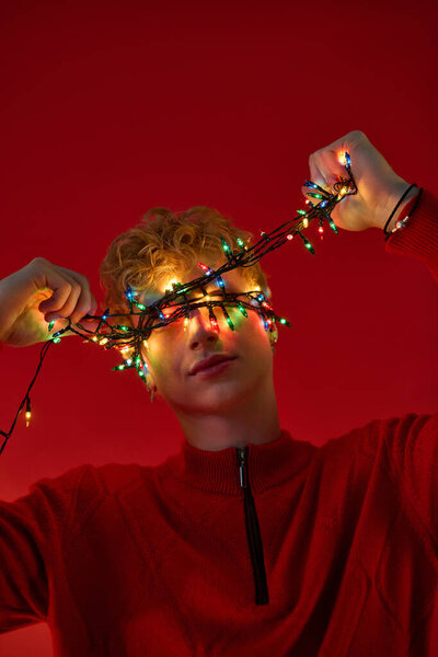 Молодой человек с вьющимися волосами игриво позирует на красном фоне, завернутый в яркие рождественские огни.